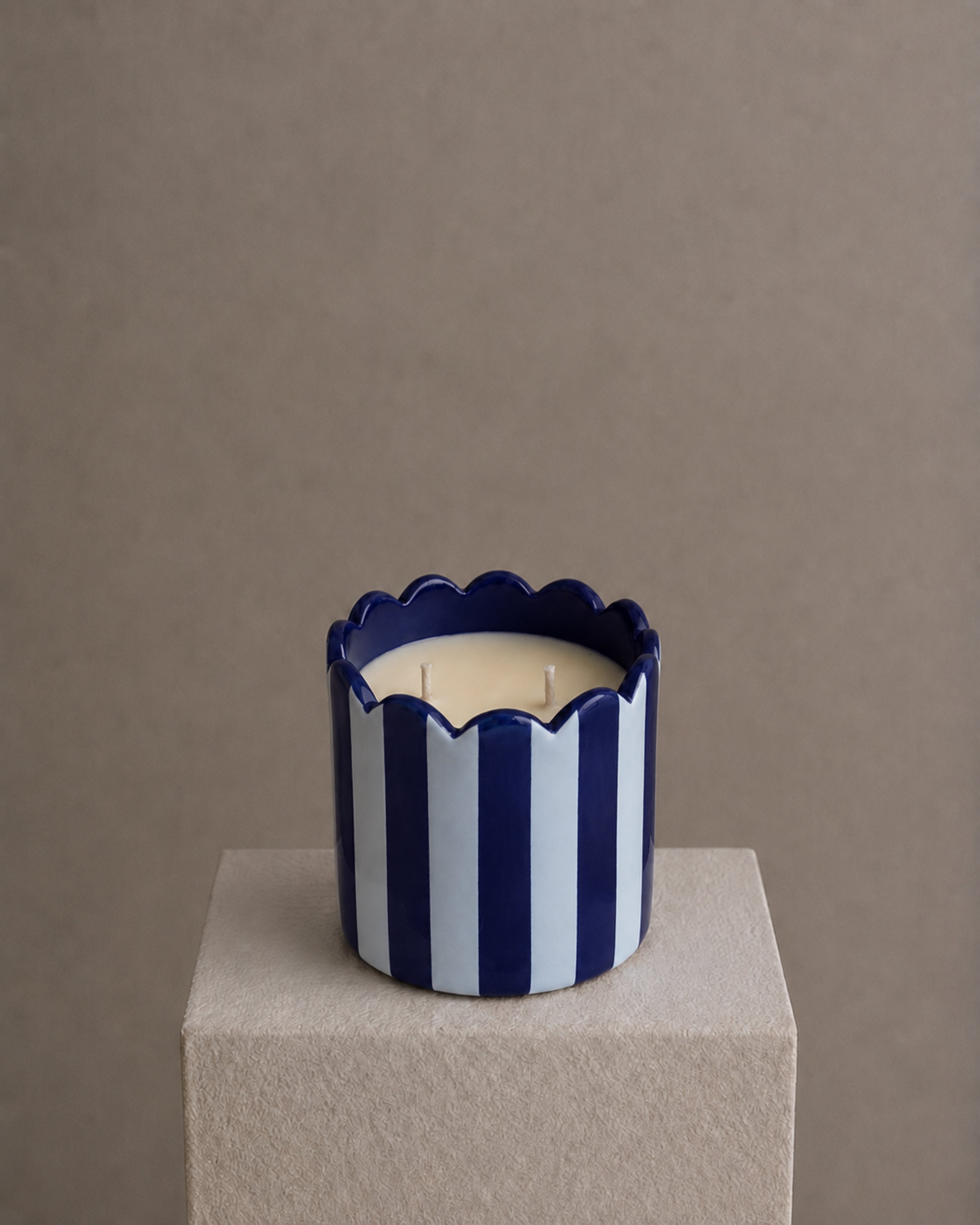A two-wick candle in a cylindrical ceramic holder with vertical navy blue and white stripes and a scalloped rim, displayed on a textured beige pedestal against a neutral taupe background.