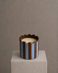 A two-wick candle in a sculptural cylindrical holder with vertical brown and light blue stripes and a scalloped rim, sitting on a textured light gray pedestal against a warm taupe background.
