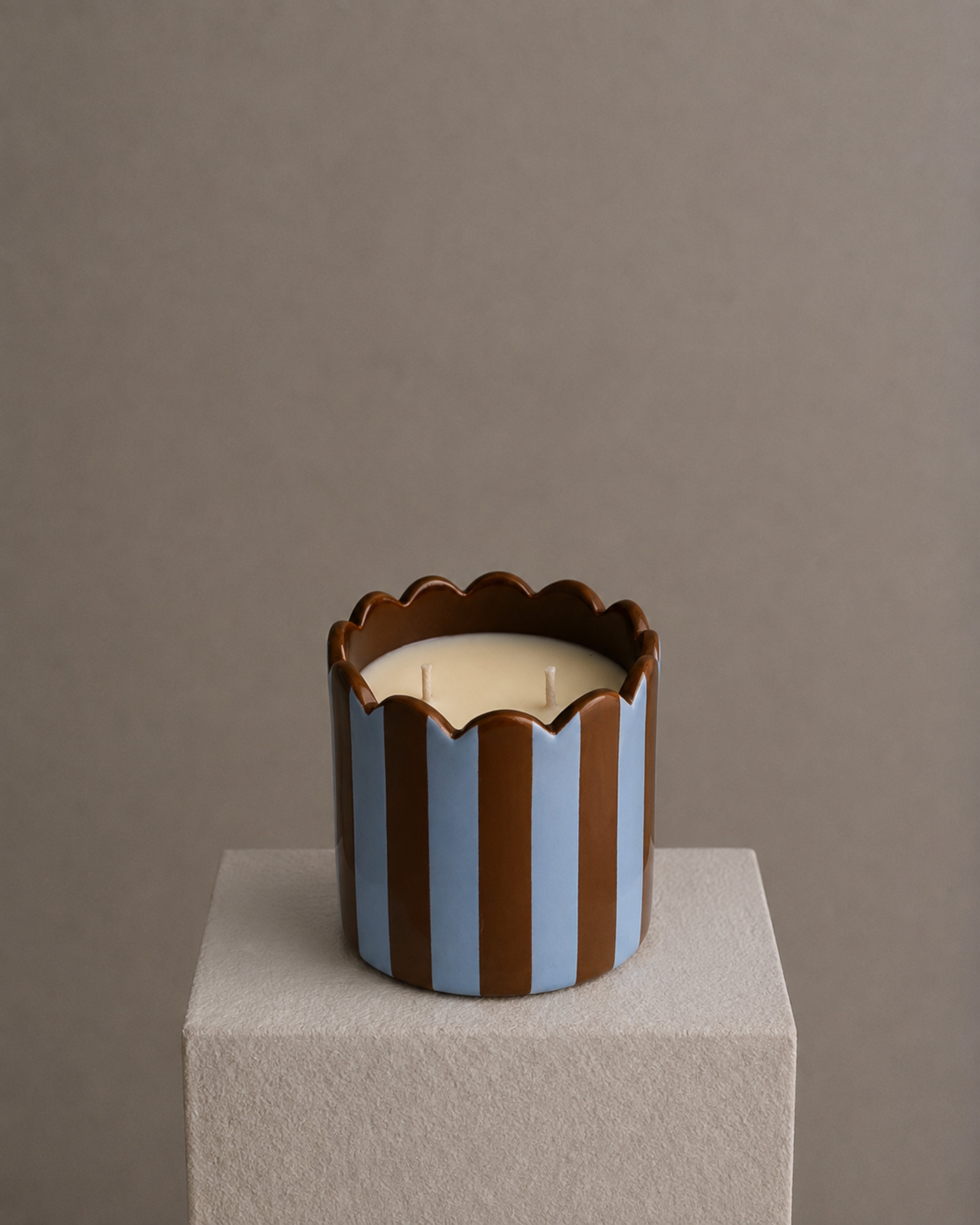 A two-wick candle in a sculptural cylindrical holder with vertical brown and light blue stripes and a scalloped rim, sitting on a textured light gray pedestal against a warm taupe background.