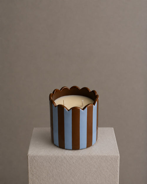 A two-wick candle in a sculptural cylindrical holder with vertical brown and light blue stripes and a scalloped rim, sitting on a textured light gray pedestal against a warm taupe background.